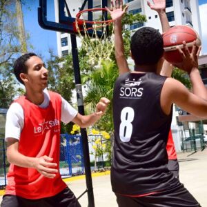 Un estudiante con camiseta roja de La Salle defiende a otro jugador con camiseta negra y el número 8, sosteniendo un balón de baloncesto en una cancha al aire libre