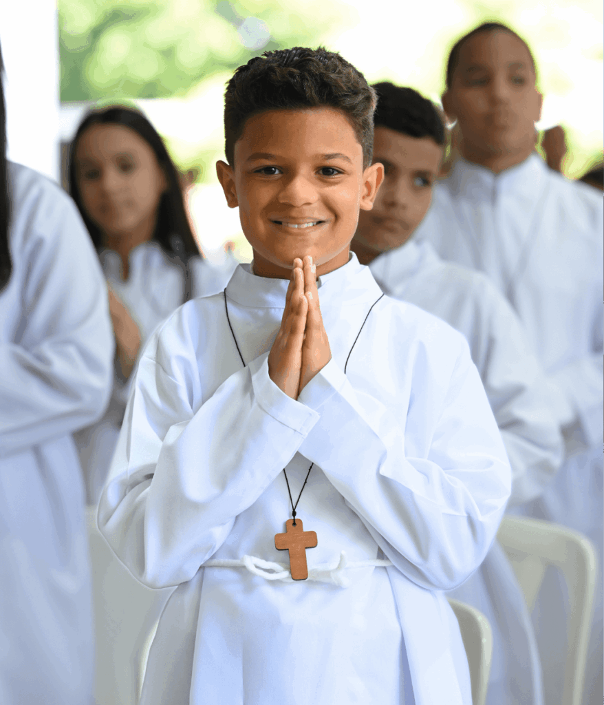 Niño del Colegio La Salle vestido de blanco sonríe con las manos juntas en señal de oración, mientras otros estudiantes aparecen al fondo en un ambiente religioso escolar.