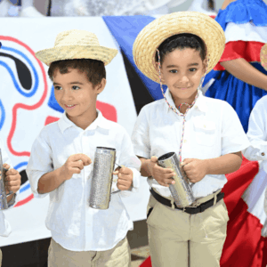 Dos niños del Colegio La Salle vestidos con ropa blanca y sombreros de paja sostienen güiras durante una actividad cultural dominicana.
