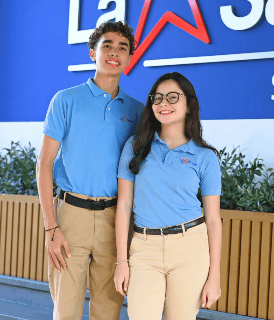Dos estudiantes del Colegio La Salle, un joven y una joven, posan sonrientes usando polo azul institucional y pantalón beige frente al logo del colegio.