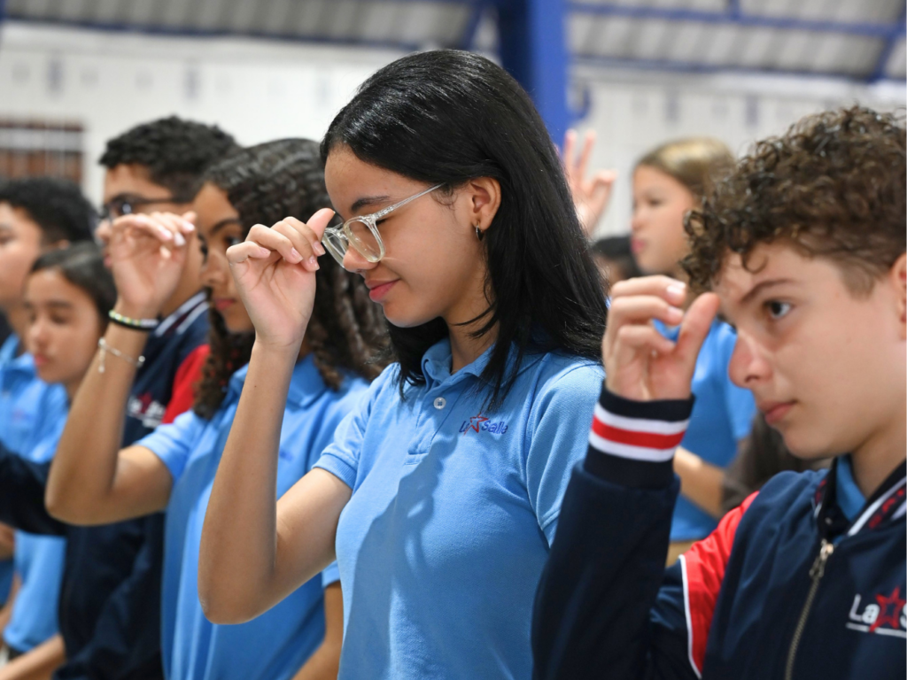 Estudiantes del Colegio La Salle con uniforme azul participan en un momento de oración dentro de un espacio escolar, con las manos levantadas en señal de fe y recogimiento.