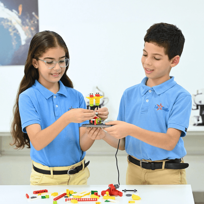 Dos estudiantes del Colegio La Salle con uniforme azul manipulan una estructura de robótica hecha con piezas tipo LEGO sobre una mesa, en un espacio educativo tecnológico.